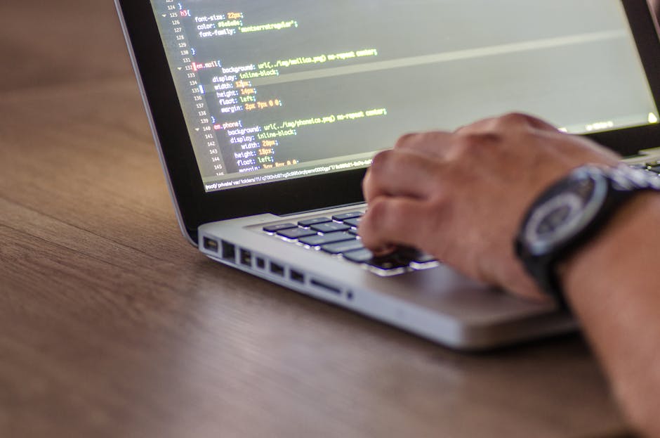 A close-up image of a person's hand wearing a wristwatch, typing on a laptop keyboard placed on a wooden desk. The laptop screen displays lines of code with various colors for syntax highlighting. The
