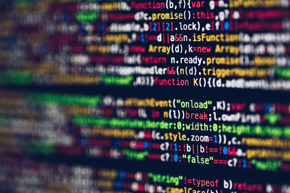 A close-up image of a person's hand wearing a wristwatch, typing on a laptop keyboard placed on a wooden desk. The laptop screen displays lines of code with various colors for syntax highlighting. The