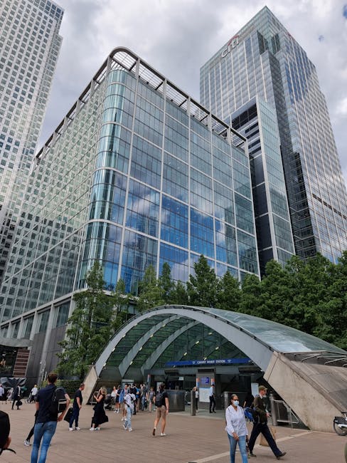 The image shows the modern exterior of a large glass office building in Canary Wharf, with reflective windows that mirror the partly cloudy sky. Adjacent to the building, there is a landscaped area wi