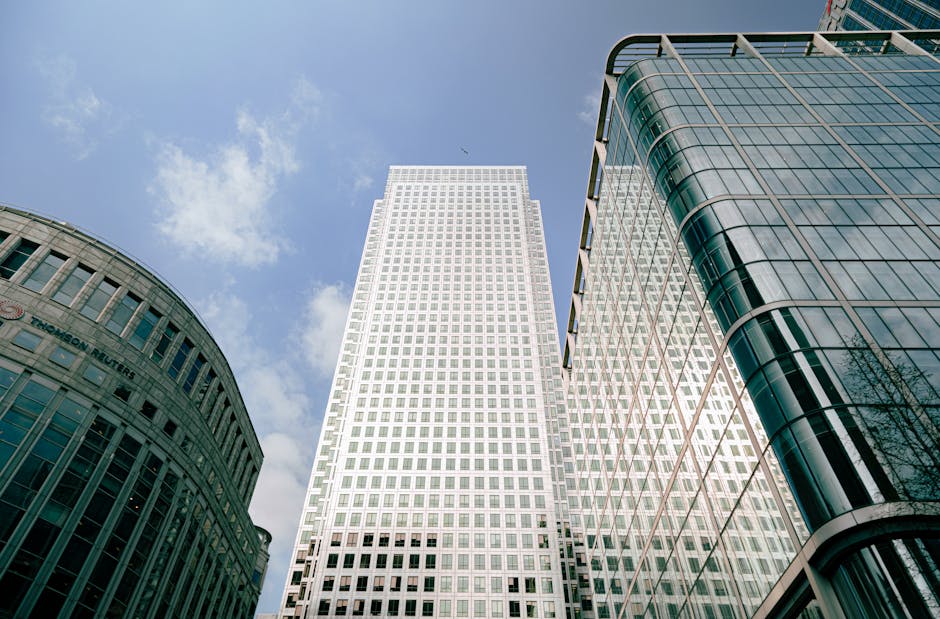 The image shows the modern exterior of a large glass office building in Canary Wharf, with reflective windows that mirror the partly cloudy sky. Adjacent to the building, there is a landscaped area wi